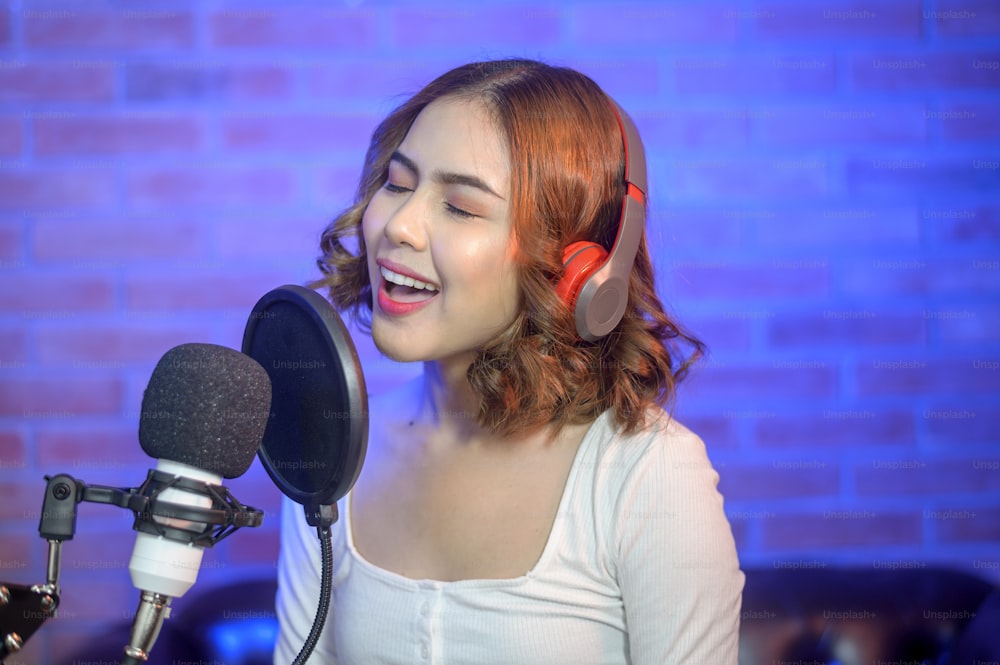 A young smiling female singer wearing headphones with a microphone while recording song in a music studio with colorful lights.