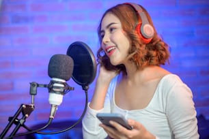 Une jeune chanteuse souriante portant des écouteurs avec un microphone tout en enregistrant une chanson dans un studio de musique avec des lumières colorées.