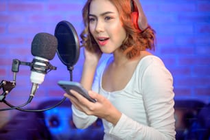 A young smiling female singer wearing headphones with a microphone while recording song in a music studio with colorful lights.