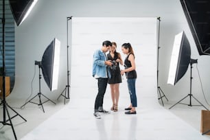 스튜디오에서 전문 사진 촬영 장면 뒤에: 아름다운 젊은 아시아 모델이 웃고 메이크업 아티스트와 함께 포즈를 취하고 있습니다. 메이크업 사진 작가가 디지털 카메라로 사진을 찍고 있습니다.