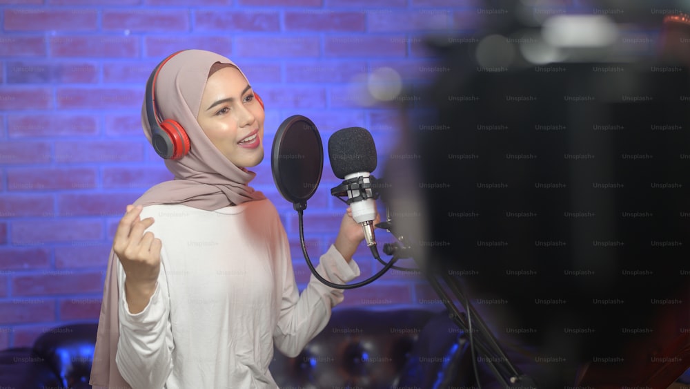 Una joven cantante musulmana sonriente con auriculares con micrófono mientras graba una canción en un estudio de música con luces de colores.