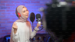 色とりどりの照明が光る音楽スタジオで、マイク付きのヘッドフォンを装着して歌を録音する若い笑顔のイスラム教徒の女性歌手。