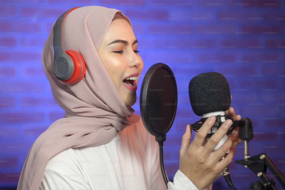 Uma jovem cantora muçulmana sorridente usando fones de ouvido com microfone enquanto gravava música em um estúdio de música com luzes coloridas.