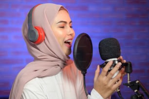 A young smiling muslim female singer wearing headphones with a microphone while recording song in a music studio with colorful lights.