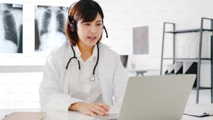 Young Asia female doctor in white medical uniform with stethoscope using computer laptop talking video conference call with patient at desk in health clinic or hospital. Consulting and therapy concept