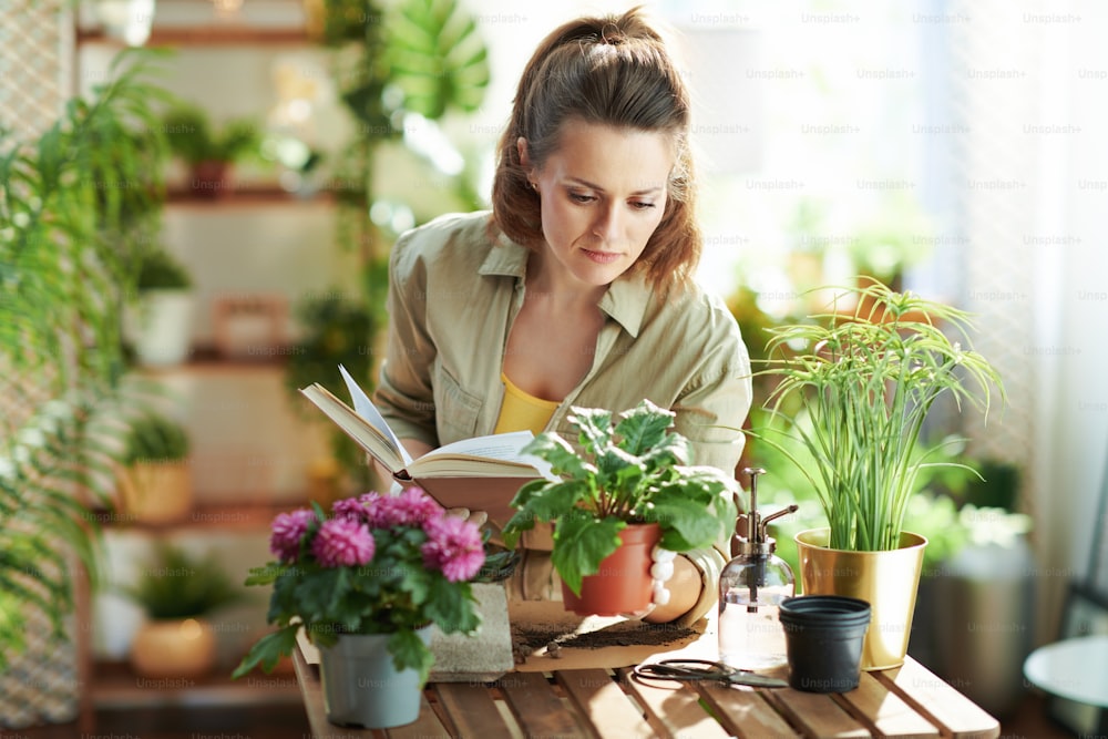 Casa verde. casalinga moderna di 40 anni in guanti di gomma bianca con pianta in vaso e libro a casa moderna in giornata di sole.