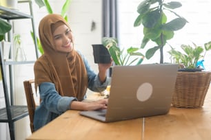 Eine junge muslimische Unternehmerin, die mit Laptop arbeitet, präsentiert Zimmerpflanzen während des Online-Livestreams zu Hause und verkauft Online-Konzept