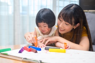 A young mom helping daughter drawing with colored pencils in living room at home.