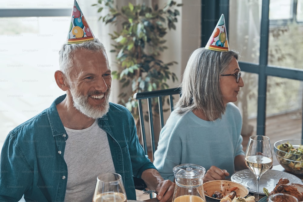 Happy senior couple in funky hats celebrating birthday while sitting at the dining table at home