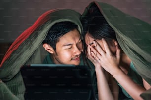 Amante adulto asiático pareja casada disfruta viendo películas de terror en línea desde una computadora portátil o tableta en manta en la cama en casa, aislamiento de cuarentena en el hogar lifetyle