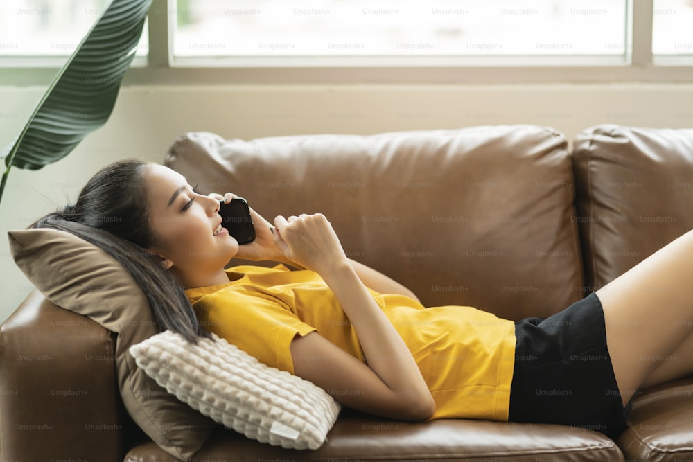 happiness cheerful smiling young adult asian female woman wear yellow cloth hand conversation on smartphone on sofa at home remote quarantine lock down period home interior background