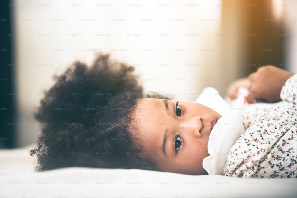 白い背景、巻き毛のアフロヘア、混合民族、かわいい女の赤ちゃん、美しい娘、家族のコンセプトで自宅のベッドに横たわったアフリカの子供のクローズアップの肖像画。幸せな女の子。