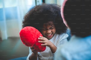 a happy little mixed afican boy holding red heart shape cushion pillow, playing with mother at home, cute boy can't hide happiness then smiling appear while with mom. family concept. white background.