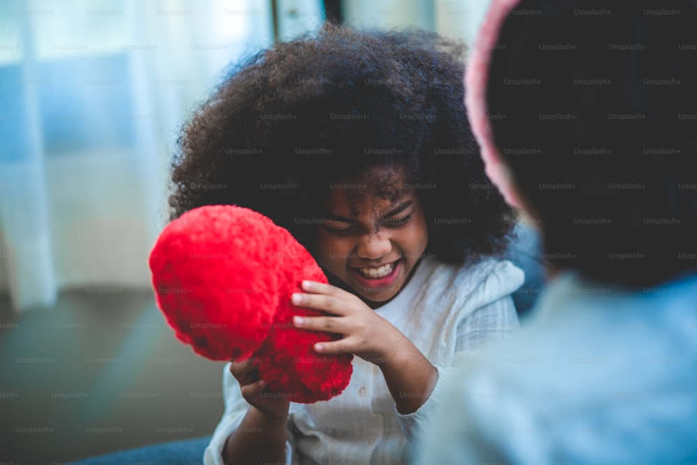 a happy little mixed afican boy holding red heart shape cushion pillow, playing with mother at home, cute boy can't hide happiness then smiling appear while with mom. family concept. white background.