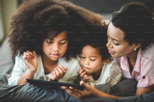 american african family spending time together at home, young boy, girl and mother. sibling using taplet, smiling kids and mom playing on the sofa bed