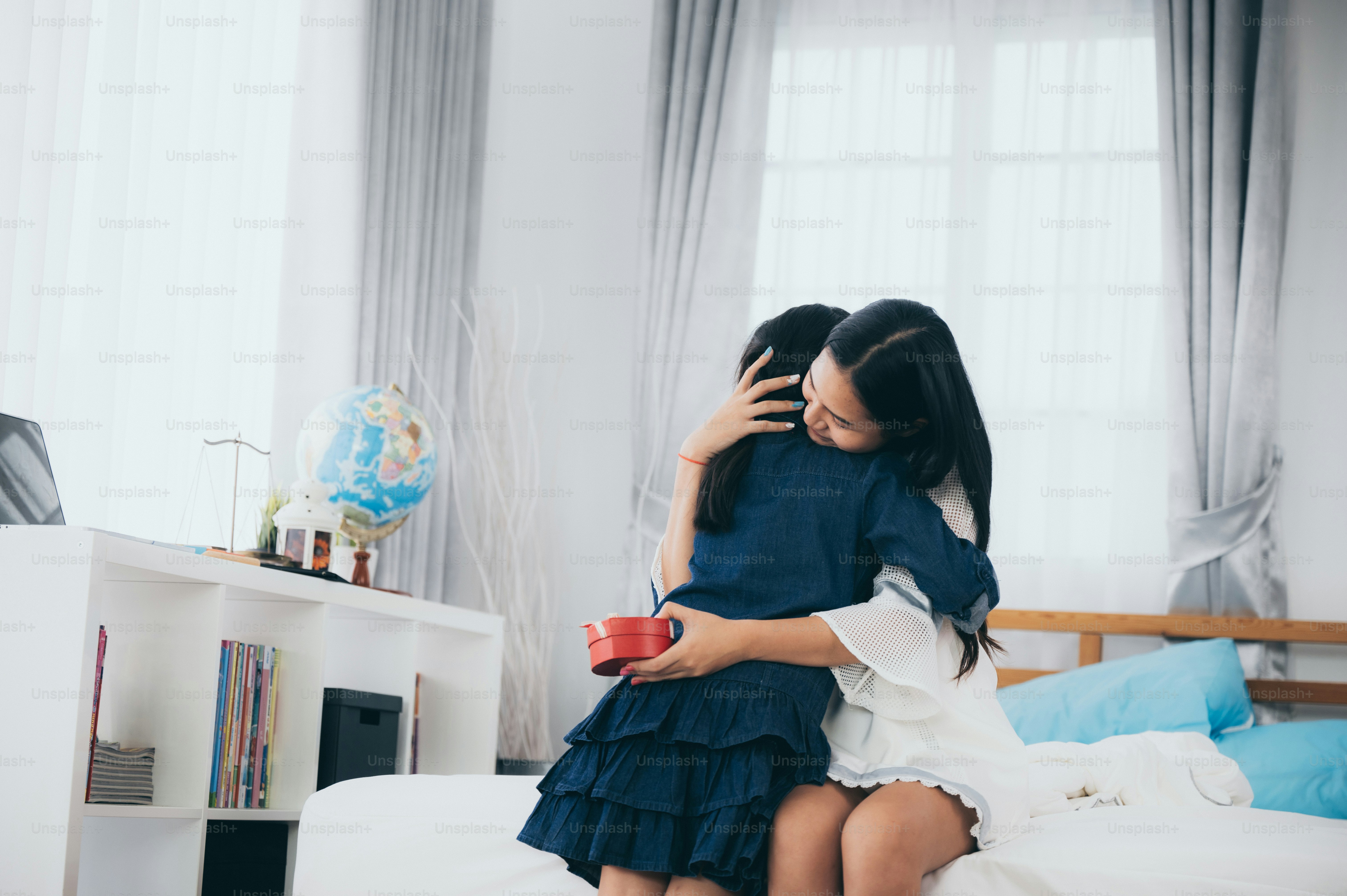 Happy mother's day! daughter surprise mom and giving her a gift box. mother hug her child, family mun and kid concept at home on the bed, asian family. kid hide present behide her back.