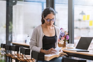 Young Asian woman wearing glasses sit and relax at the coffee shop read a book put a laptop at the table.