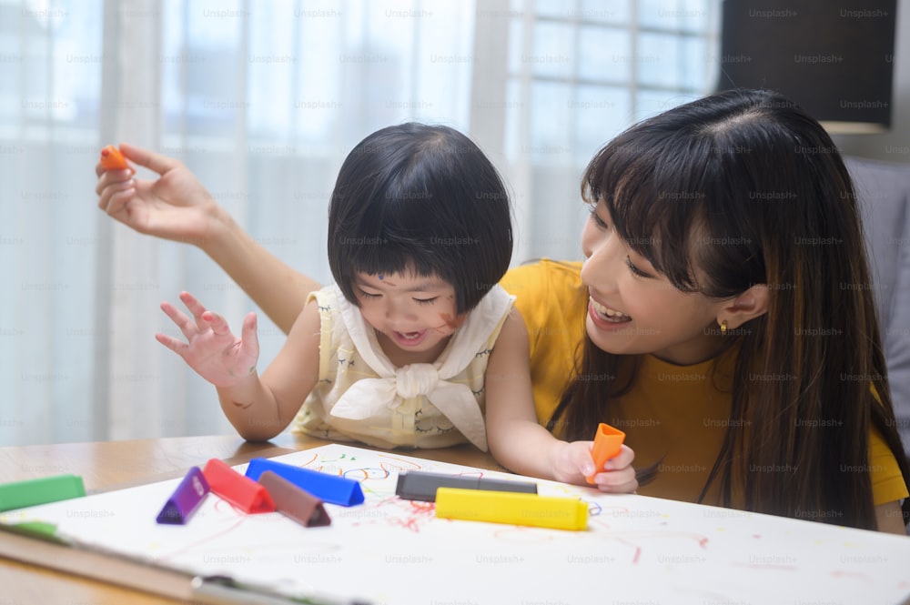 Una joven madre ayudando a su hija a dibujar con lápices de colores en la sala de estar de su casa.