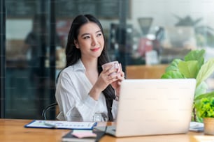 Hermosa joven empresaria asiática sonriendo sosteniendo una taza de café y una computadora portátil trabajando en la oficina.