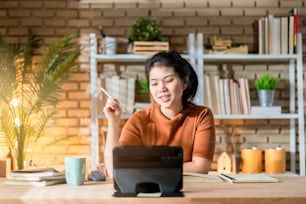 魅力的なスマートなアジアの女性は、タブレット技術デバイスを使用して自宅でカジュアルな布を着用、アジアの女性は本棚の背景を持つオンラインのホームスタジオオフィスで働いています