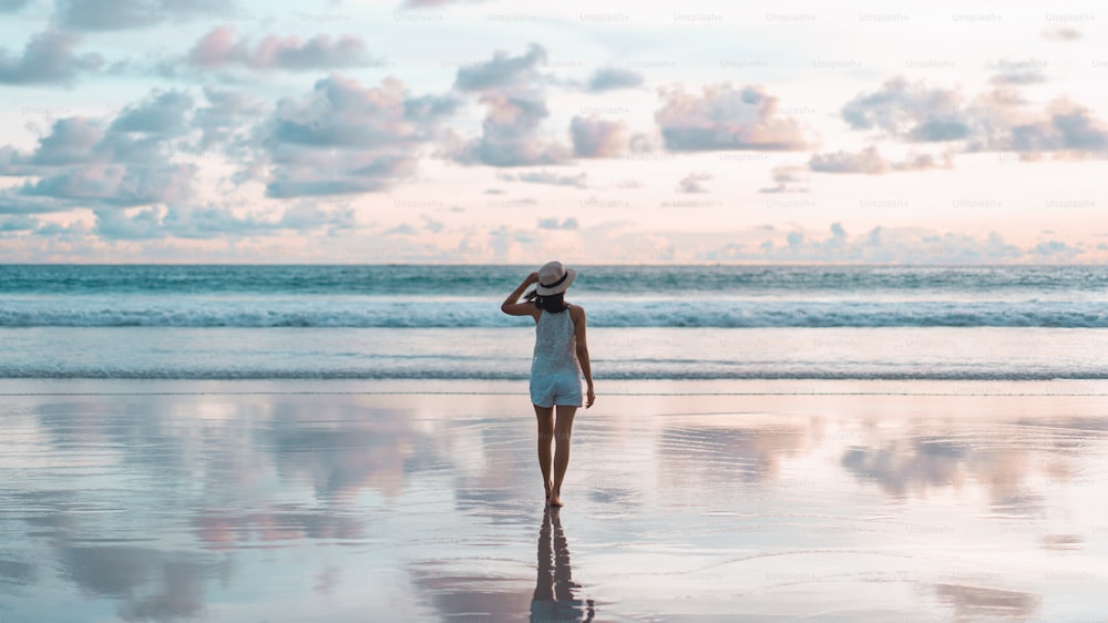 Rear view of young adult tourist asian woman walking relax on beach sand with beautiful dramatic sunset sky. Outdoor domestic travel at andaman ocean. Phuket, Thailand.