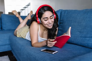Joven mujer latina leyendo libros y escuchando música con auriculares en el sofá en casa en México América Latina