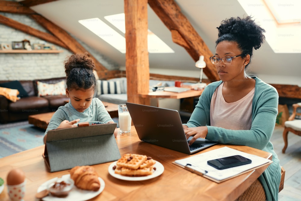 Pequeña niña negra desayunando y usando una tableta digital mientras su madre está trabajando en una computadora portátil en la mesa del comedor.