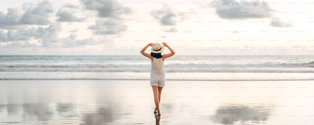 Rear view of young adult tourist asian woman walking relax on beach sand with beautiful dramatic sunset sky. Outdoor travel banner size background.