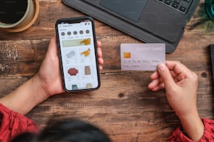 Cerrar la mano de la mujer usando la tarjeta de crédito que compra en línea en la aplicación móvil en la mesa de madera en casa
