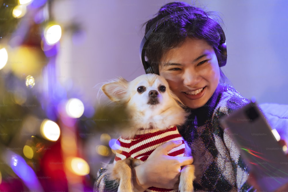 Estilo de vida de la felicidad con el perro animal, mujer asiática sonriente amistosa sostiene al pequeño perrito faldero mientras escucha música auriculares wnjoy navidad año nuevo weelend vacaciones en la noche celebrándose a sí misma