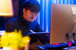 young Asia freelance creative designer programer focus working hand using pen with tablet thinking concentrate working late at home overtime at night low light ambient in living room with city bokeh