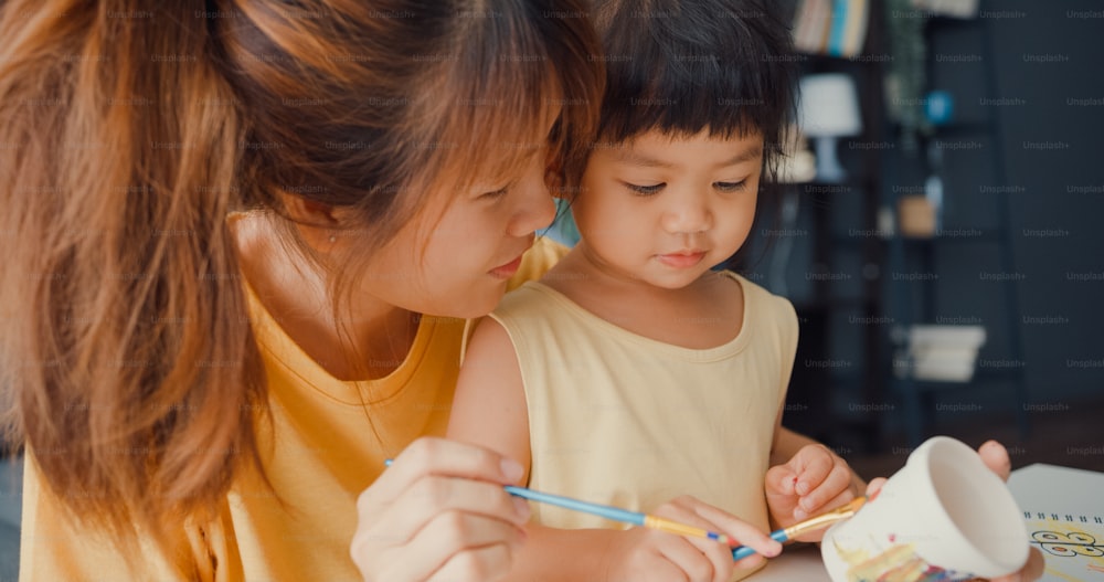 Mamá de la familia Happy Asia enseña a una niña pequeña a pintar la olla de cerámica divirtiéndose relajándose en la mesa de la sala de estar de la casa. Pasar tiempo juntos, Distancia social, Cuarentena para la prevención del coronavirus.