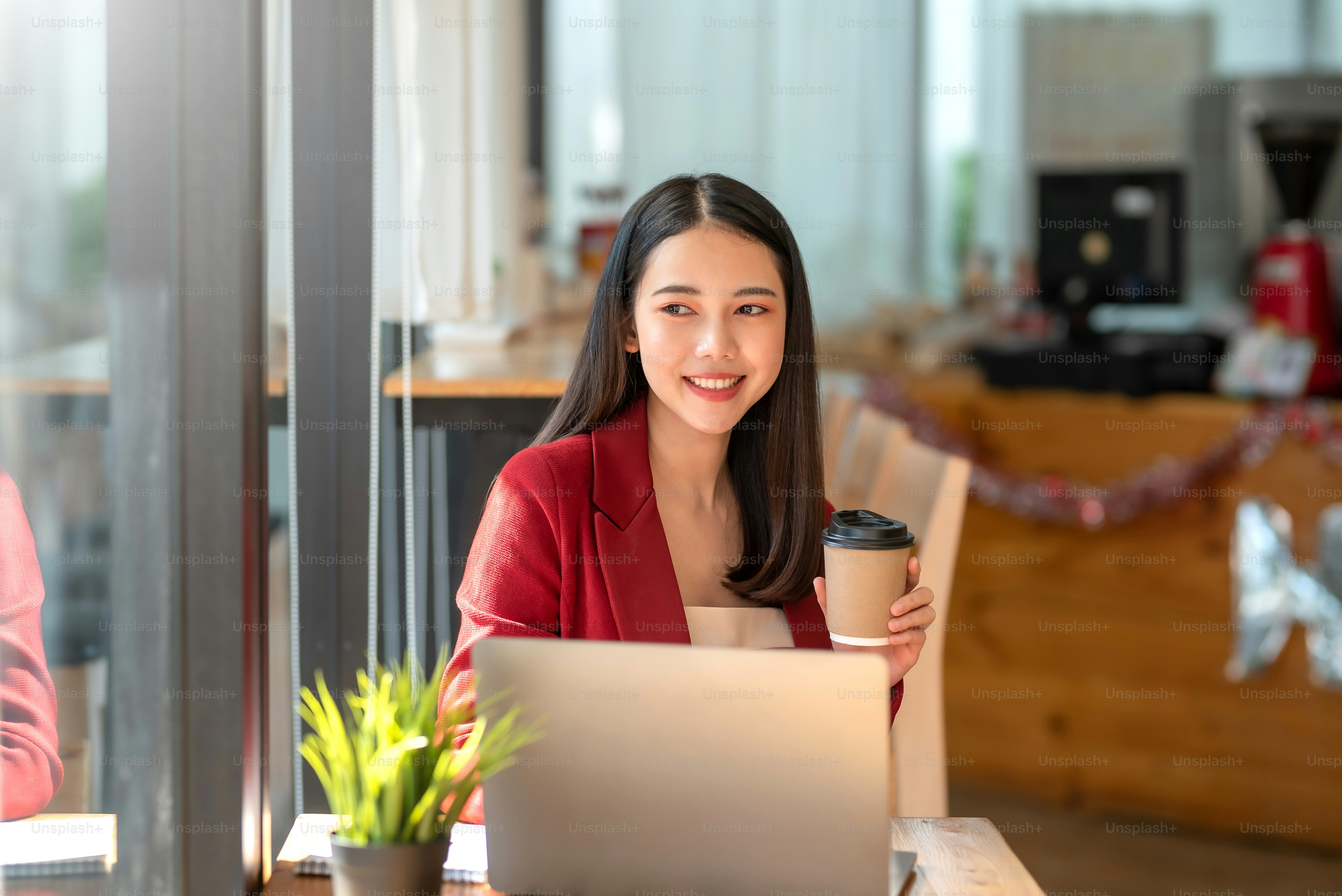 Glückliches Lächeln sitzt junge asiatische Geschäftsfrau im roten Anzug mit Kaffee und Laptop im Café.