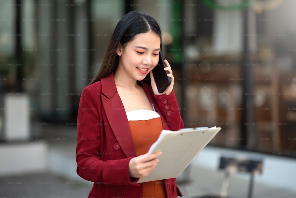Confident Asian businesswoman in a red suit standing with document talking on the phone with her client outside the office.