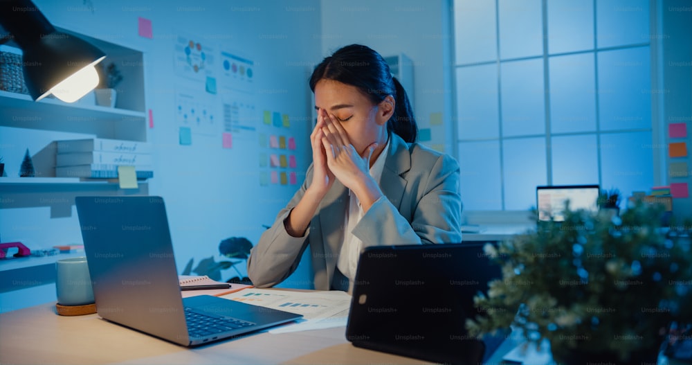 La giovane donna d'affari asiatica si siede con laptop e tablet sulla scrivania strofinando l'occhio sente dolore e stanca dal superlavoro in ufficio di notte. Le donne soffrono di sindrome dell'ufficio a lungo termine, salute mentale sul posto di lavoro.