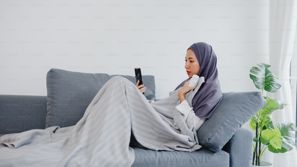 Joven mujer musulmana de Asia usa hiyab usando videollamada telefónica hablando con consulta médica o consulta en línea en el sofá en la sala de estar de casa. Distanciamiento social, cuarentena por concepto de coronavirus.