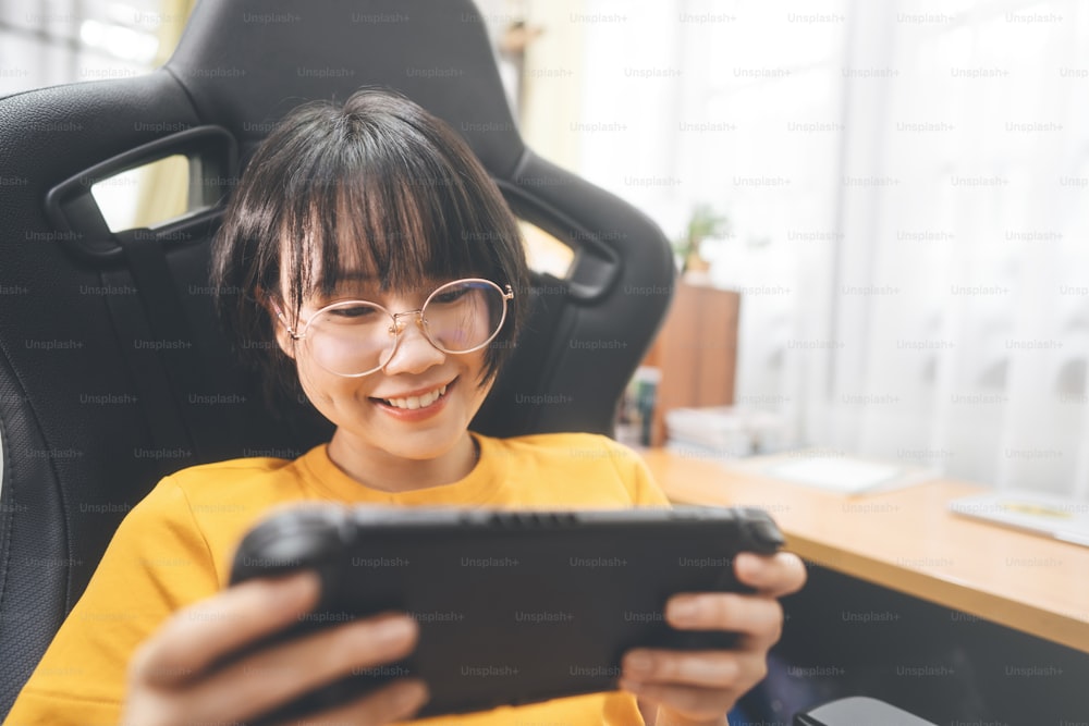 Happy smile nerd young adult asian gamer woman wear eyeglasses play a online game on handheld portable device. People leisure lifestyle at home.