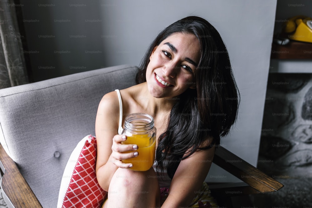 メキシコラテンアメリカの自宅でソファで休んでいる間、オレンジジュースを飲むラテン系の女性