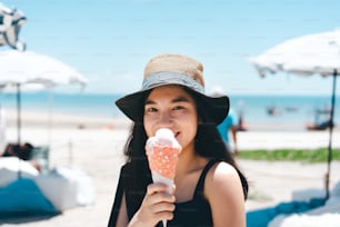 夏休みのコンセプトで旅行。若い大人のアジアの女性は、ビーチの海の屋外カフェでリラックスします。日光の下でアイスクリームコーンを食べる幸せな笑顔。