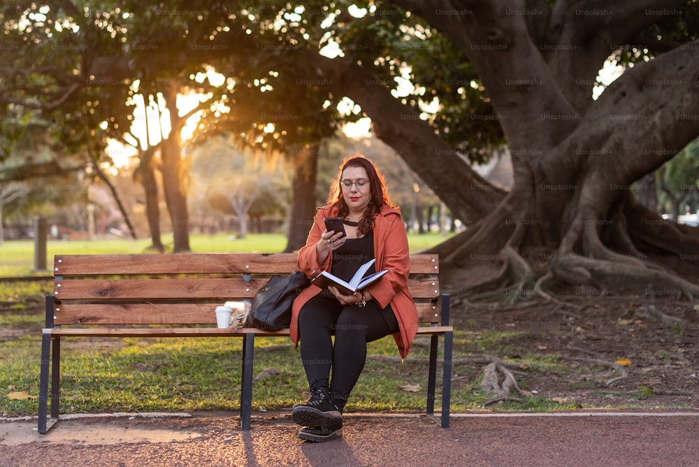 屋外で働くエグゼクティブプラスサイズの女性。彼女は公園のベンチに座りながらノートを持ち、テキストメッセージを送っています