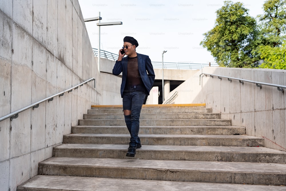 Young Latin gay man wearing a fashionable hat and sunglasses, walking down the stairway while texting on his smartphone, isolated on a white background. LGTB. Copy space