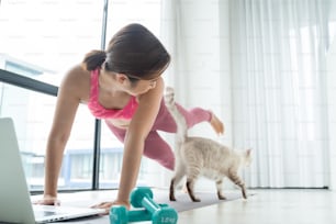若いアジアの美しい女性は、猫と一緒に自宅でフィットネスをトレーニングし、ラップトップでオンラインチュートリアルの練習トレーニングフィットネスワークショップを見ています。