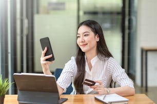 Mulher asiática jovem bonita segurando smartphone e usando o cartão de crédito para compras on-line.