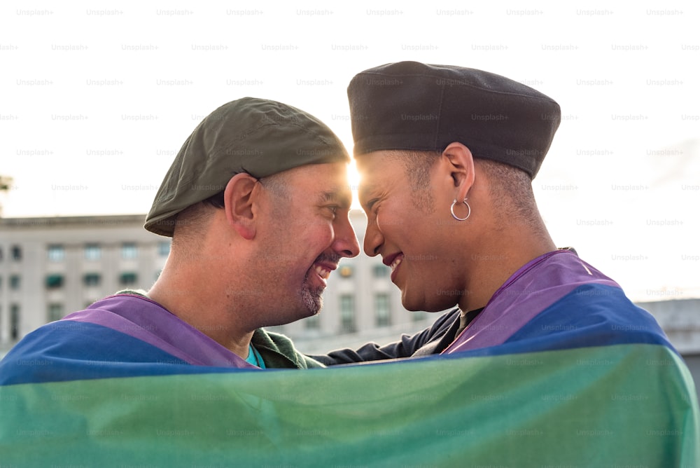 LGBTのレインボーフラッグの下で、笑いながらお互いを見つめ合うゲイカップル。プライドのコンセプト。プライド月間。