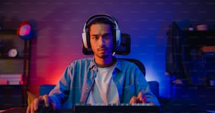 幸せなアジアの男ゲーマーはヘッドフォン参加を身に着けて、夜のリビングルームでビデオゲームカラフルなネオンライトコンピューターをプレイします。オンラインのeスポーツストリーミングゲーム、自宅検疫活動のコンセプト。
