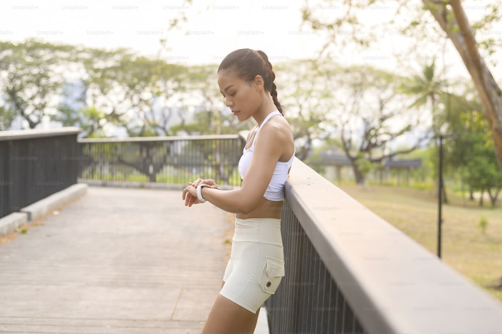 都市公園で運動しながらスマートウォッチを使用するスポーツウェアを着た若いフィットネス女性、健康とライフスタイル。