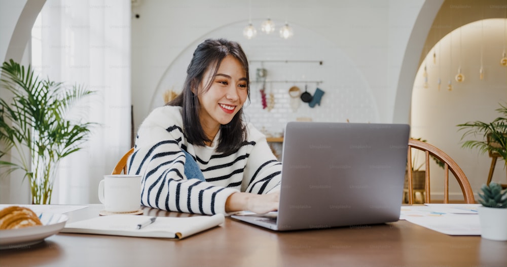 젊은 아시아 여성 프리랜서는 집에서 부엌에서 회사와의 원격 회의를 위한 계정 및 재무 프로젝트를 위해 노트북으로 프레젠테이션 작업에 중점을 둡니다. 여학생은 집에서 온라인으로 학습합니다.