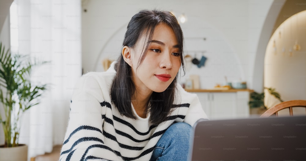 Young Asian lady freelance focus on presentation work in laptop for account and finance project for remote meeting with company in kitchen at house. Girl student learn online at home.