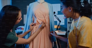 캐주얼 초과 근무를 공유하는 두 명의 전문 아시아 쾌활한 여성 패션 디자이너 팀워크는 밤에 가게에서 더미에 아름다운 드레스를 만듭니다. 레이디 재단사 비즈니스 파트너 창업 사업.