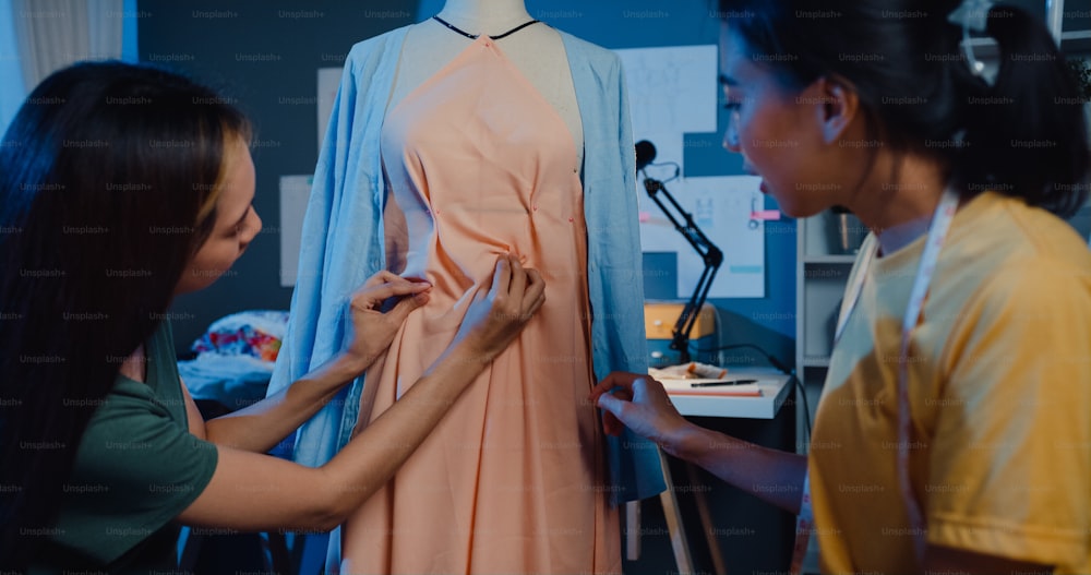 Dos profesionales de Asia alegre diseñadora de moda femenina trabajan en equipo con el trabajo casual de horas extras comparten la idea de crear un hermoso vestido en el maniquí en la tienda por la noche. Dama sastre socia comercial de inicio de negocio.